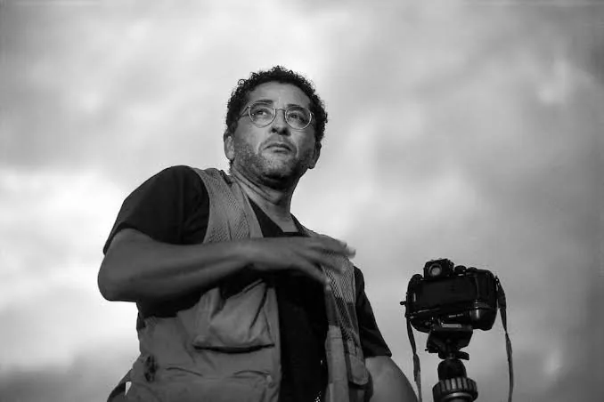 Araquém Alcântara em retrato em preto e branco, ao lado de uma câmera sobre tripé, sob um céu nublado